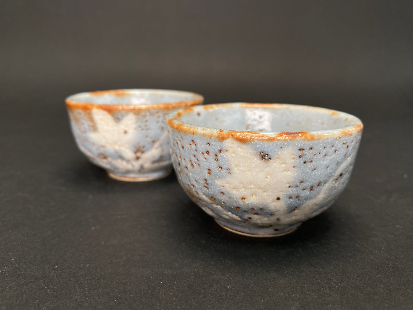 Pair of handmade Nezumi Shino Mino ware sake cups with blue-grey glaze, reddish-brown rims, rust iron spots, and abstract white plant patterns