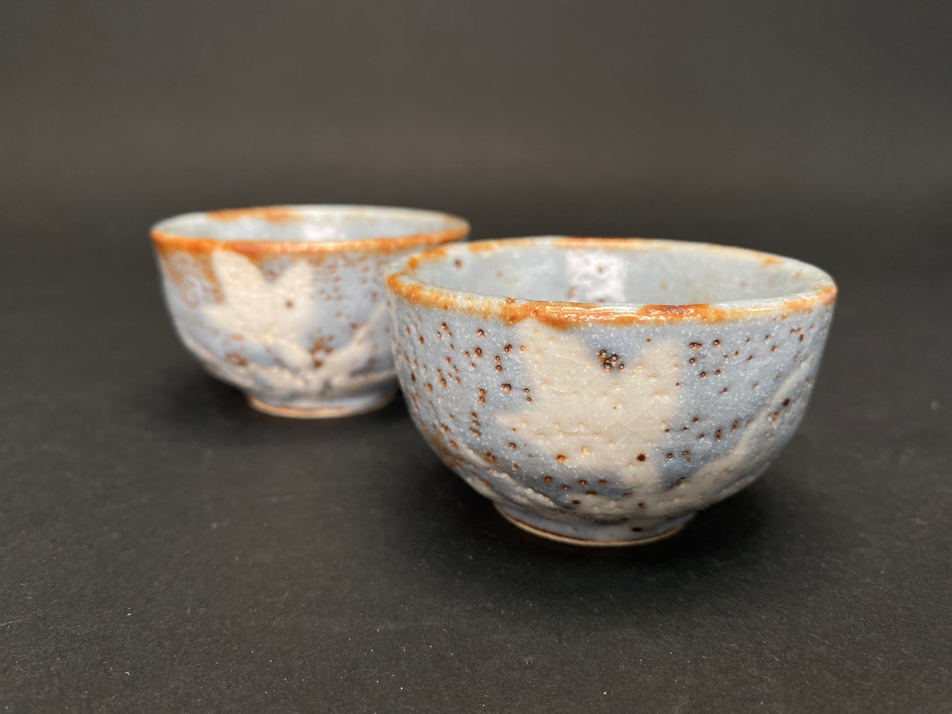 Pair of handmade Nezumi Shino Mino ware sake cups with blue-grey glaze, reddish-brown rims, rust iron spots, and abstract white plant patterns
